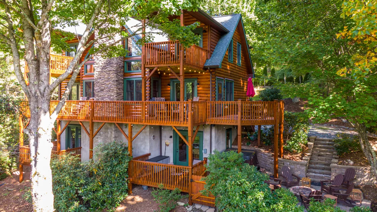 The image showcases a classic log cabin nestled in lush greenery, featuring expansive wooden balconies perfect for soaking in mountain sunsets. An inviting outdoor seating area sits nearby, ideal for stargazing or gathering around the fire pit with friends and family.
