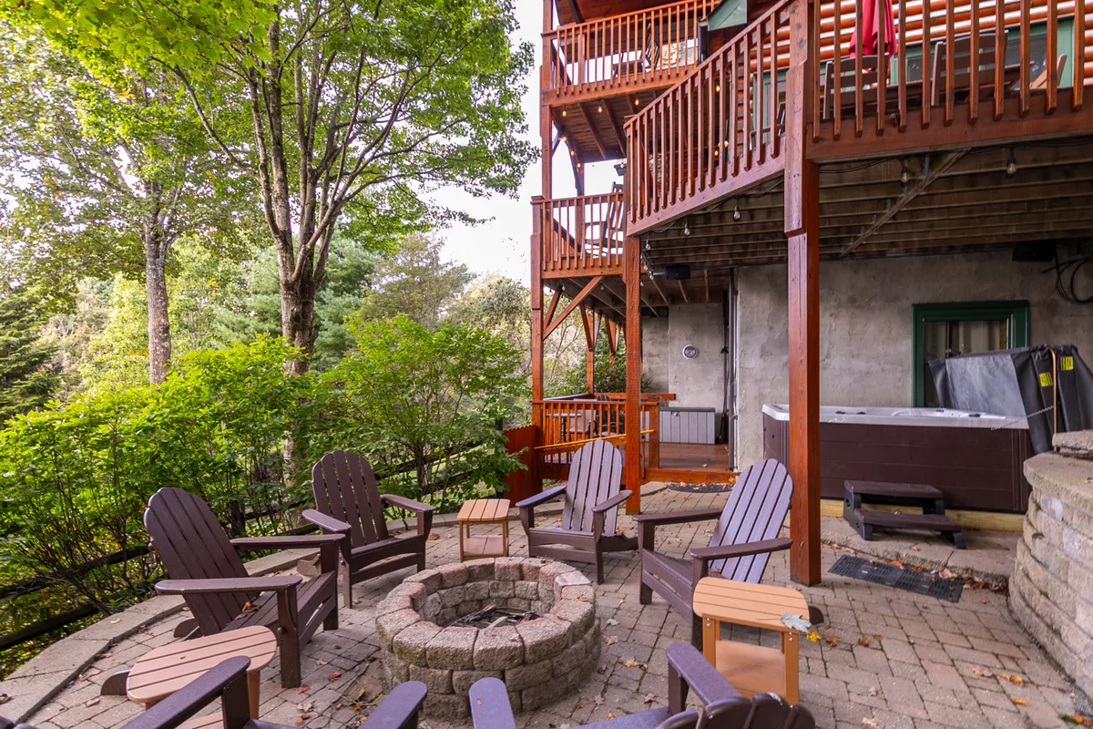 This exterior image captures a cozy fire pit surrounded by adirondack chairs and a bubbling hot tub on a wooden deck. Guests are seen gathering around the fire, relaxing in the hot tub, and enjoying outdoor dining in a communal, mountain retreat setting.