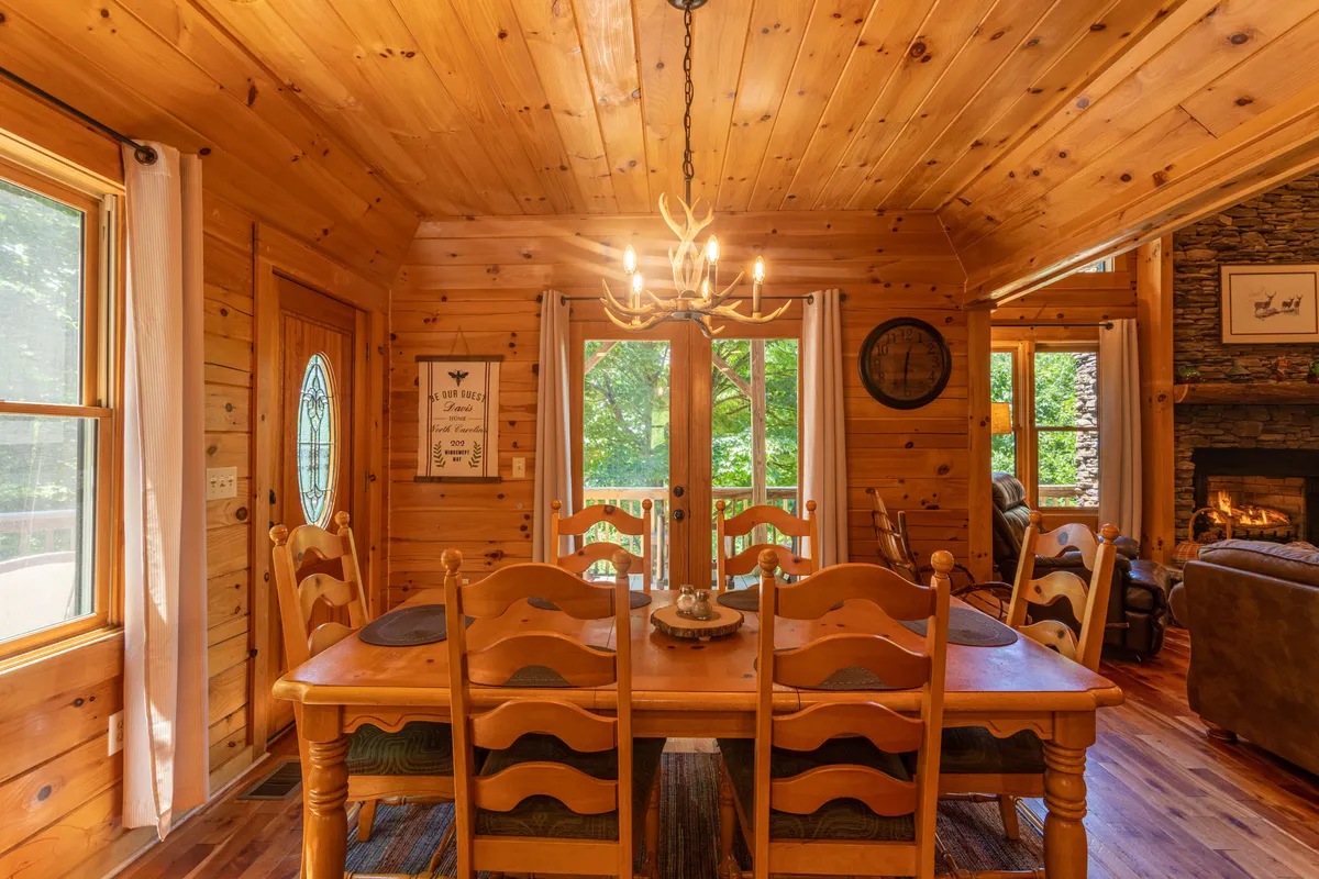 An open-plan dining area featuring a large wooden table beneath a stylish chandelier, backed by floor-to-ceiling windows and a fireplace, with wooden walls adding to the rustic charm.