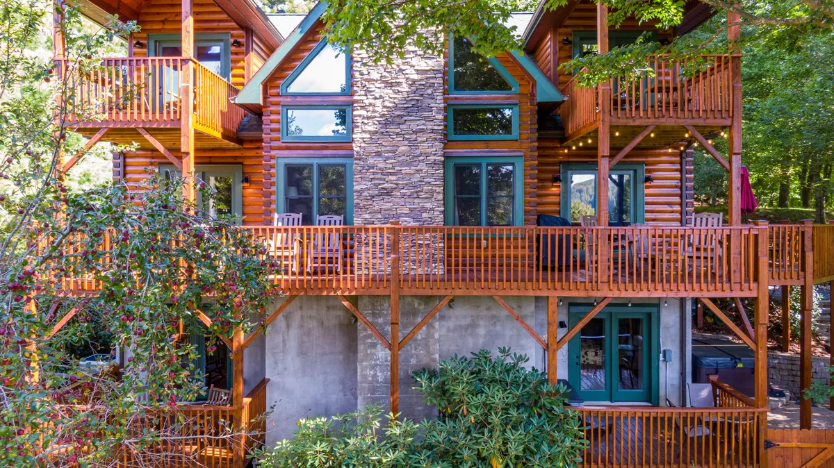 The exterior of the cabin is showcased with wooden balconies, a stone facade, and large windows, all nestled amidst lush greenery. This angle highlights the property’s inviting presence and its seamless blend with the surrounding landscape.