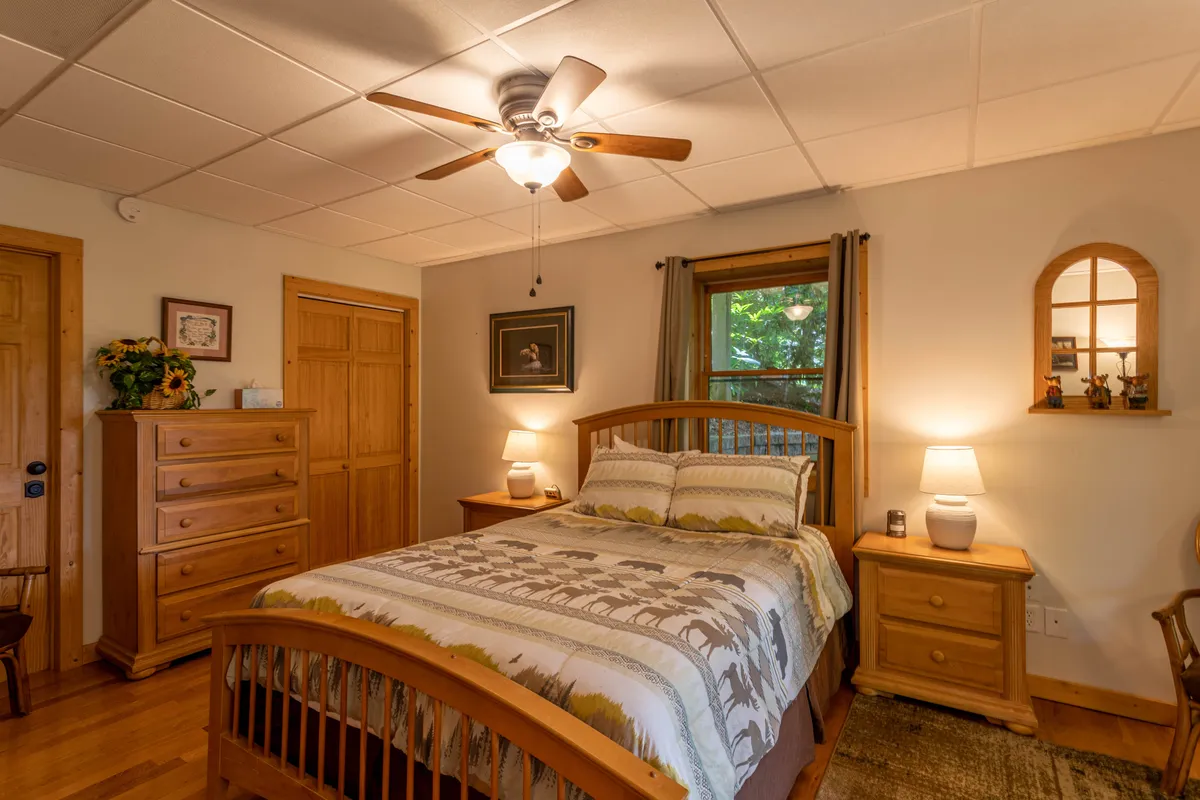 A cozy bedroom with a bed featuring patterned bedding, a wooden dresser, framed artwork on the wall, and a window allowing natural light to pour in. The scene is warm and inviting, perfect for restful moments and quiet relaxation.
