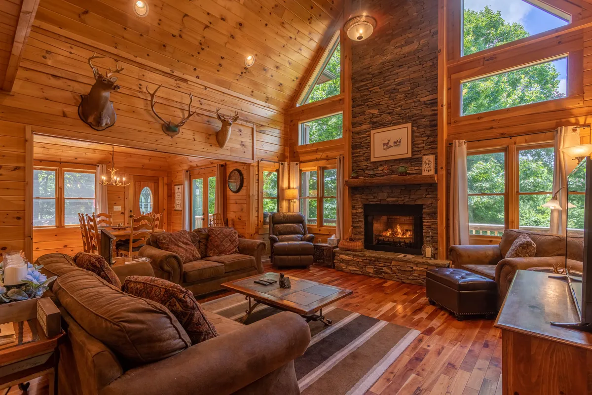 A spacious living room with a towering stacked stone fireplace, large windows revealing sweeping mountain views, wooden ceiling beams, antler decor, and plush, comfortable seating arranged for social gatherings.