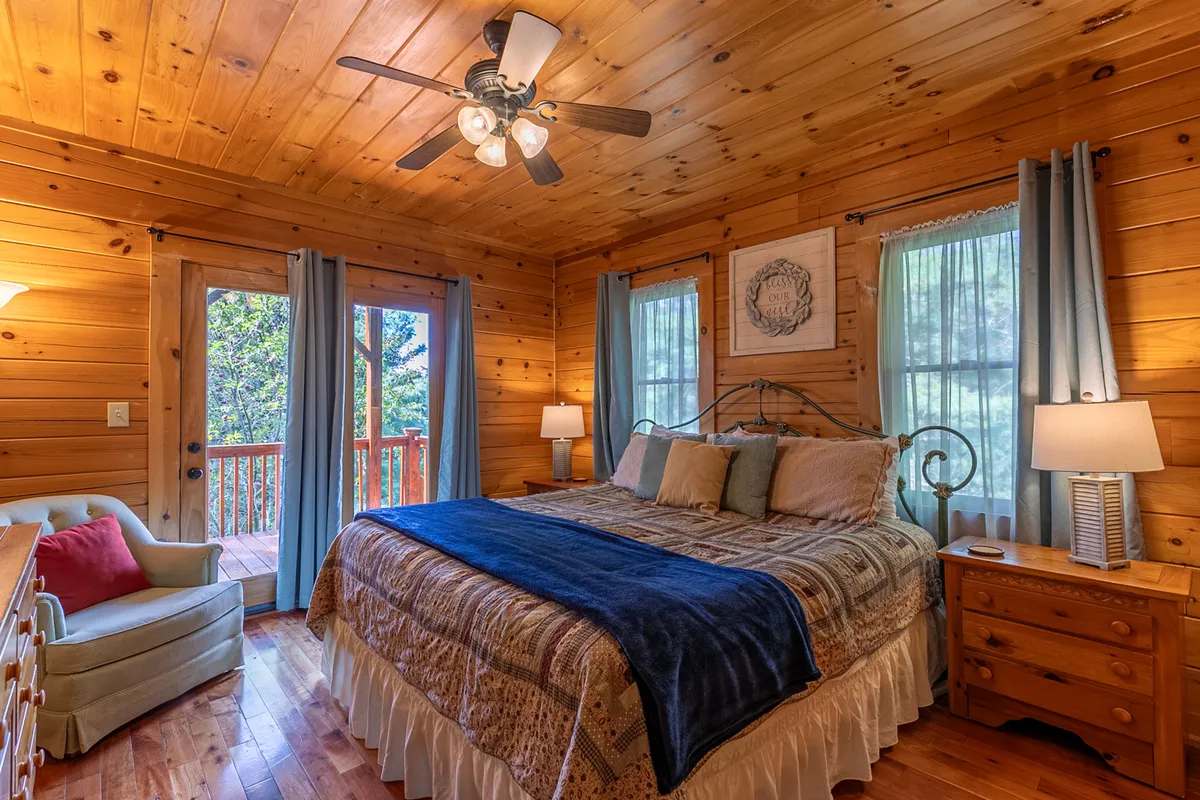 A tranquil bedroom with a comfortable bed, large windows offering serene views of nature, a dresser, and a plush armchair. The space radiates peace and is ideal for unwinding or enjoying a quiet morning coffee.
