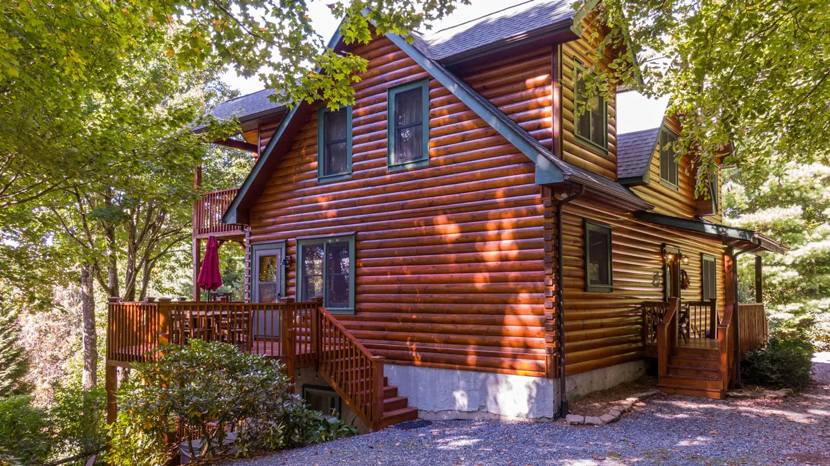 An inviting exterior view of the log cabin featuring a spacious wooden deck that opens onto lush greenery, suggesting the presence of a large, secure yard. The scene includes families gathering on the deck, surrounded by nature, with ample space for pets and children to play nearby.