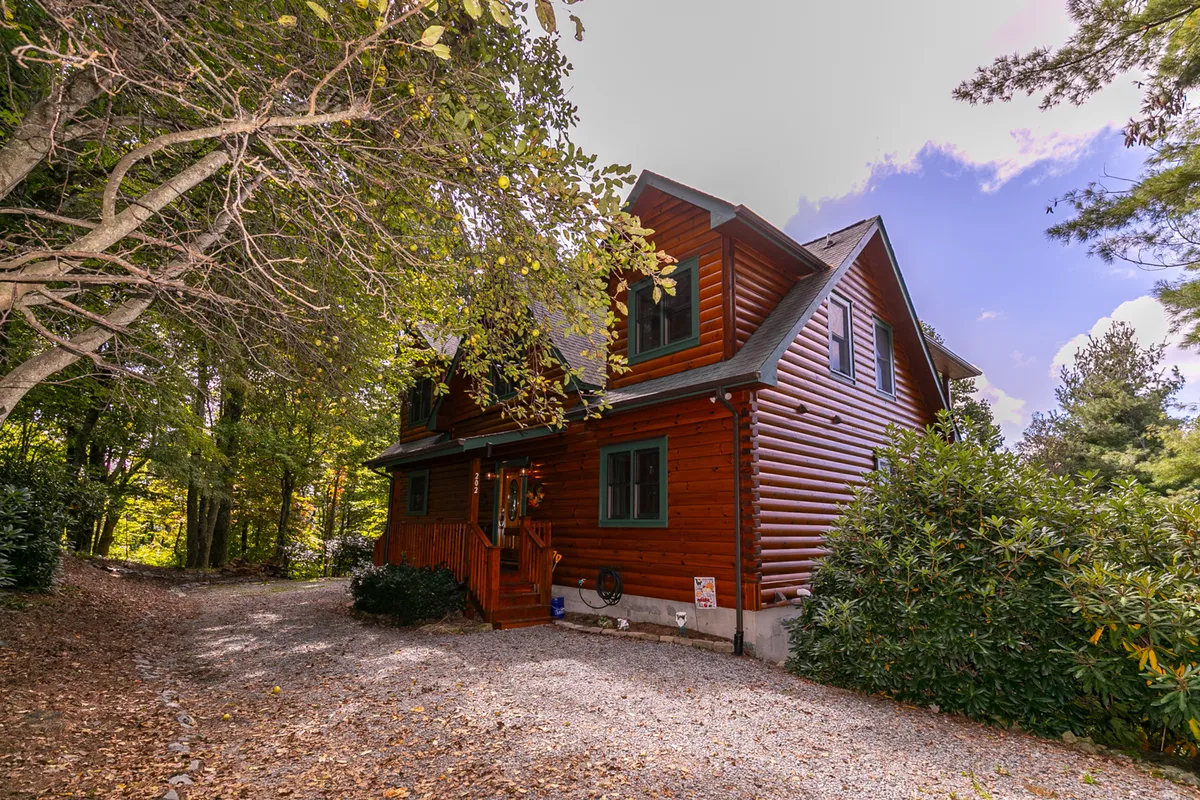 The image features the log cabin’s exterior, including inviting wooden front steps, mature trees, and a gravel driveway. The setting feels private and is enveloped in nature, highlighting the mountain retreat’s connection to the outdoors and potential nearby walking areas.
