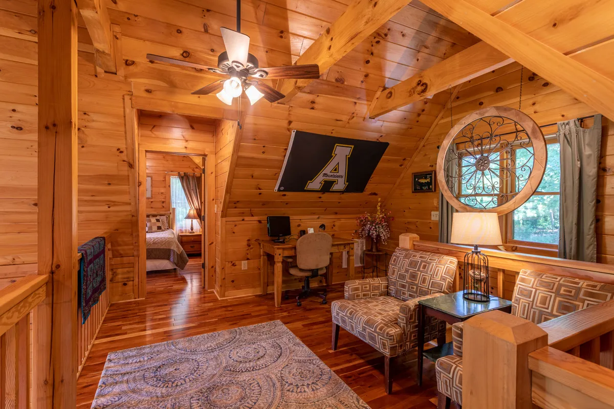 A warm and welcoming loft area featuring wooden beams, a comfortable sofa seating area, a distinctive circular window, and a desk with a chair. The space is cozy, bright, and lends itself both to relaxation and focus.