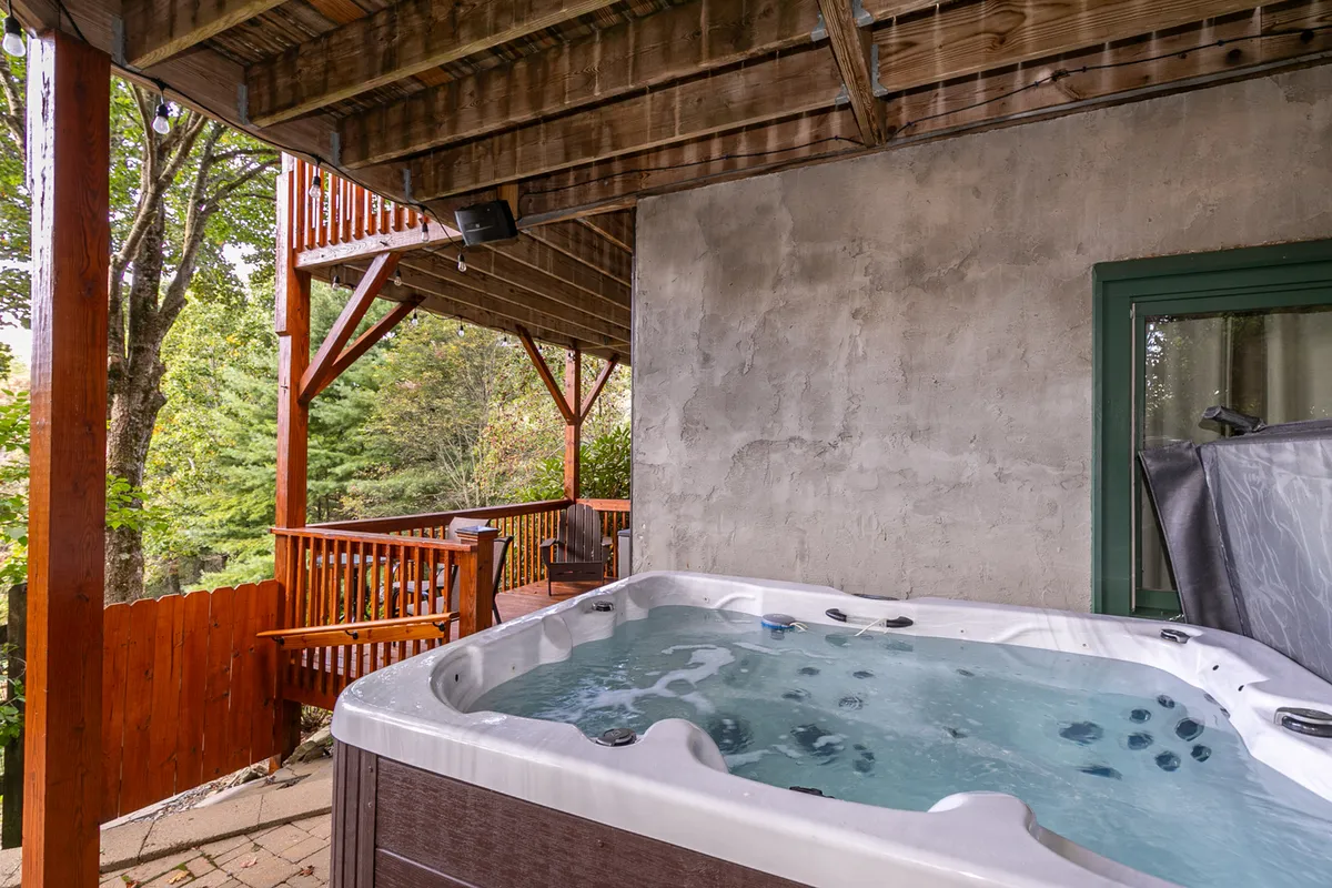A tranquil exterior deck scene showcasing a bubbling hot tub surrounded by wooden railings and tall trees. The cozy setup offers privacy while immersing guests in natural beauty and is ideal for relaxing after active days exploring the mountains.