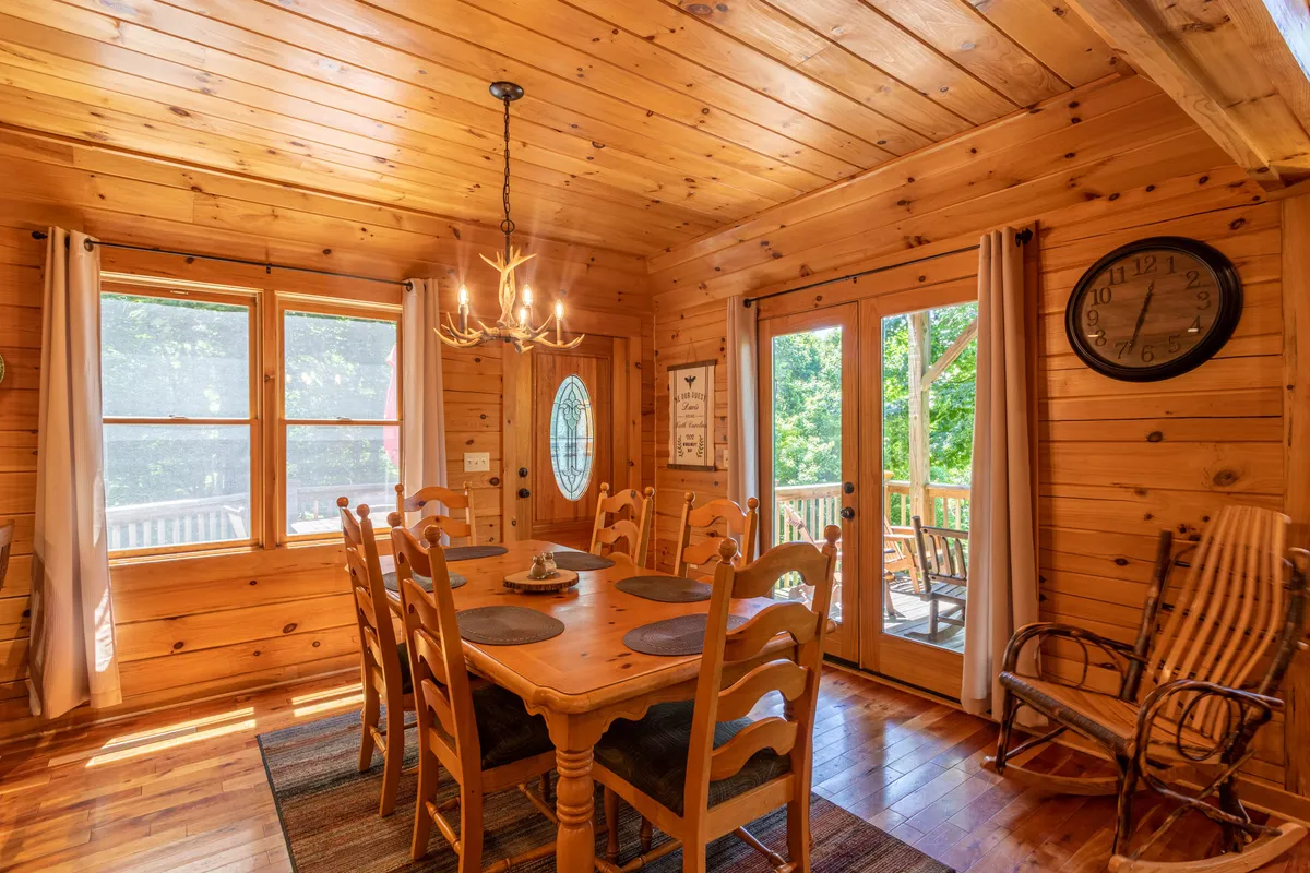 A cozy and light-filled dining area with a visible door leading outside, surrounded by windows that offer views of the greenery and yard. The setting features a large dining table, warm wood accents, and a sense of easy movement between indoor living spaces and the yard.