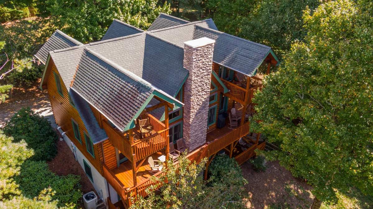 This image shows a spacious wooden deck attached to the cabin, with a prominent stone chimney and surrounded by lush green foliage. The deck is equipped for relaxation and offers space for outdoor gatherings, with ample room for pets to roam while owners enjoy the view.