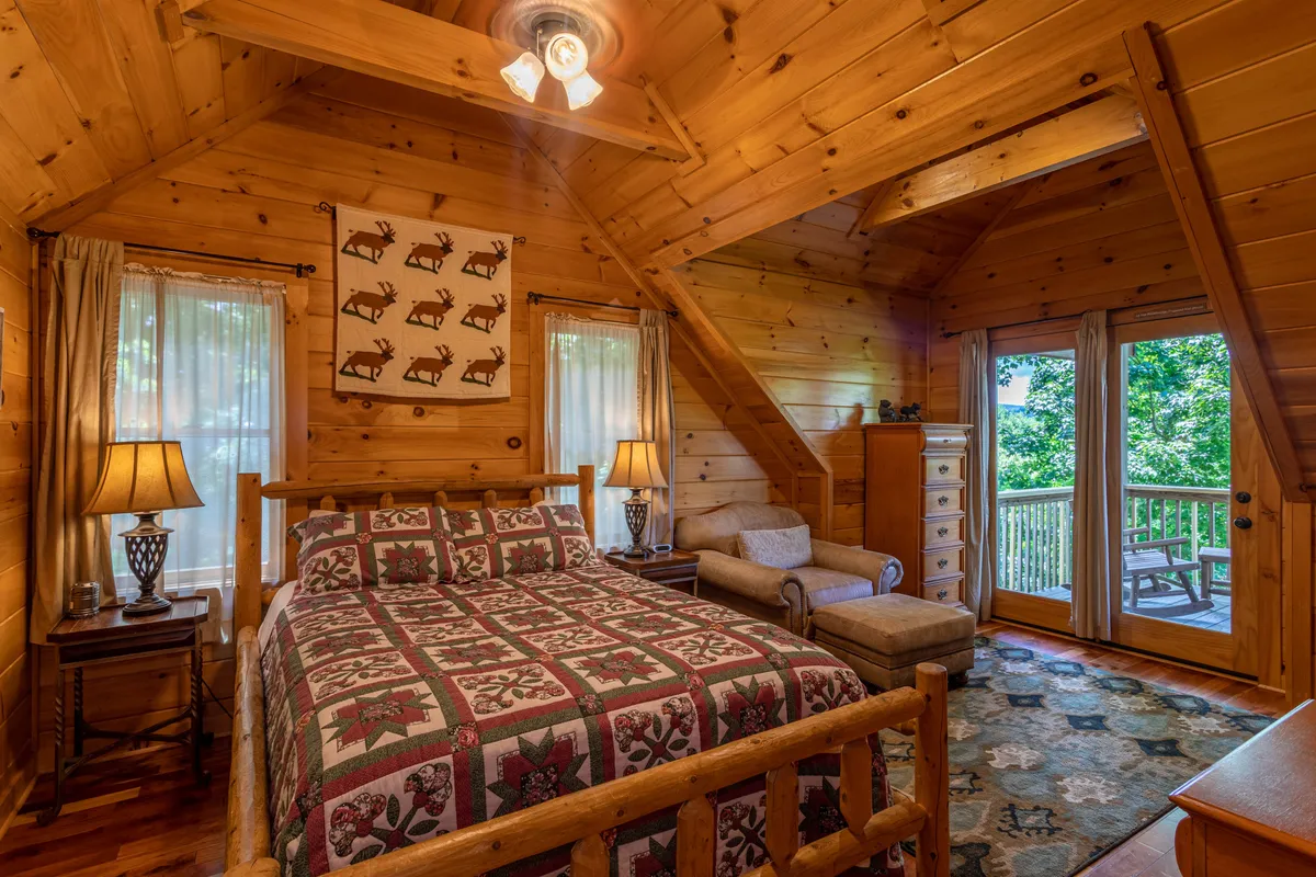 A warm bedroom with a wooden bed, plaid quilt, lamps, and large windows opening onto a private deck, offering direct access to and views of nature. The setting is comfortable and inviting, ideal for quiet moments or taking breaks while enjoying the landscape.