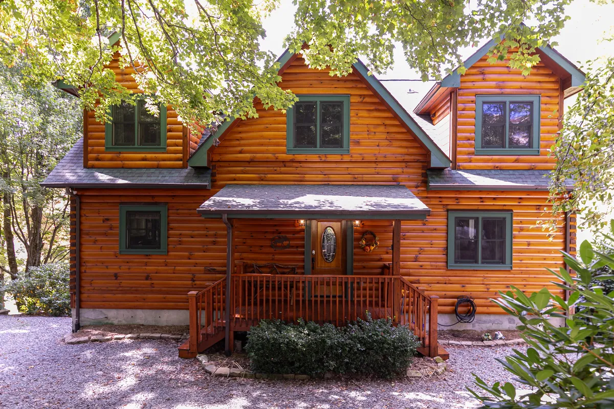 A classic log cabin exterior with a welcoming front porch, green window accents, and dense woods in the background, setting the stage for mountain adventures. The scene hints at the home’s proximity to nature and the possibilities for outdoor exploration just steps away.